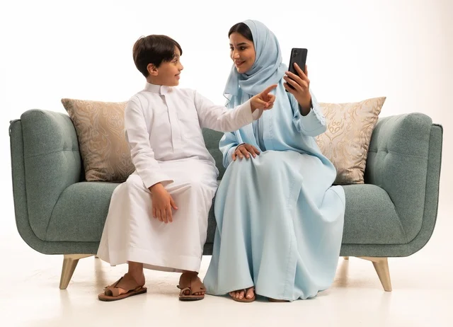 الاجتماع الأسري في غرفة المعيشة وبناء مجتمع سعودي متنامي, التقاط سيلفي عائلي تذكاري عبر الهاتف المحمول, بورتريه لسيدة عربية سعودية خليجية مبتسمة تقوم بتوثيق اللحظات الممتعة بإيماءات السعادة والفرح مع ابنها, فرحة الأبناء بقضاء وقت ممتع مع الأهل, خلفية بيضاء