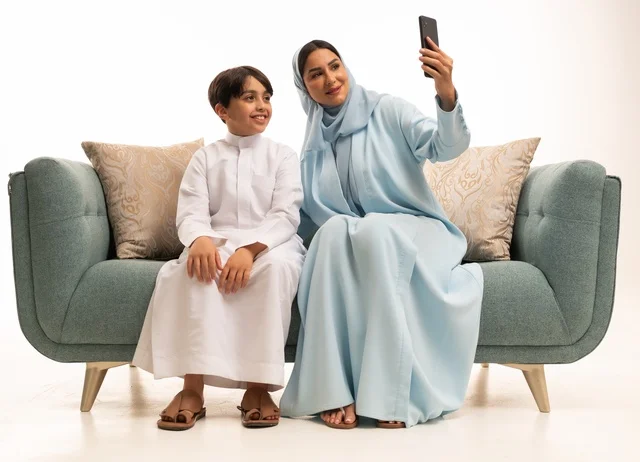 الاجتماع الأسري في غرفة المعيشة وبناء مجتمع سعودي متنامي, التقاط سيلفي عائلي تذكاري عبر الهاتف المحمول, بورتريه لسيدة عربية سعودية خليجية مبتسمة تقوم بتوثيق اللحظات الممتعة بإيماءات السعادة والفرح مع ابنها, فرحة الأبناء بقضاء وقت ممتع مع الأهل, خلفية بيضاء