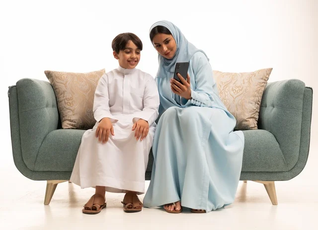 The family gathering in the living room and building a growing Saudi community, taking a memorable family selfie via mobile phone, a portrait of a smiling Arab Saudi Gulf woman documenting enjoyable moments with gestures of happiness and joy with her son, the joy of children spending quality time with family, white background.