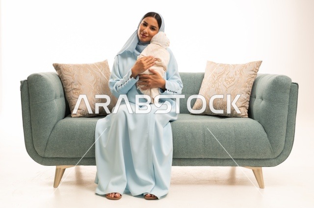 مساعدة الرضع على الاسترخاء والهدوء والنوم، تقوية رابط الأمومة بين الأم وطفلها، مفهوم البراءة والطفولة، تحمل المسؤولية والعناية بالأطفال،  بورتريه لإمرأة عربية خليجية سعودية محجبة تجلس على الاريكة تحمل صغيرها بإيماءات الحب والحنان, خلفية بيضاء
