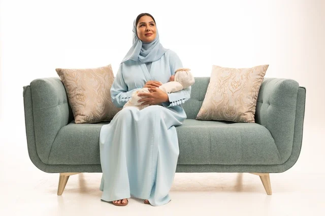 مساعدة الرضع على الاسترخاء والهدوء والنوم، تقوية رابط الأمومة بين الأم وطفلها، مفهوم البراءة والطفولة، تحمل المسؤولية والعناية بالأطفال،  بورتريه لإمرأة عربية خليجية سعودية محجبة تجلس على الاريكة تحمل صغيرها بإيماءات الحب والحنان, خلفية بيضاء