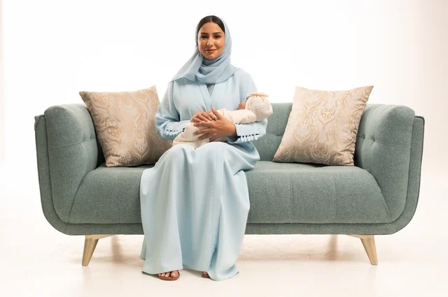 مساعدة الرضع على الاسترخاء والهدوء والنوم، تقوية رابط الأمومة بين الأم وطفلها، مفهوم البراءة والطفولة، تحمل المسؤولية والعناية بالأطفال،  بورتريه لإمرأة عربية خليجية سعودية محجبة تجلس على الاريكة تحمل صغيرها بإيماءات الحب والحنان, خلفية بيضاء