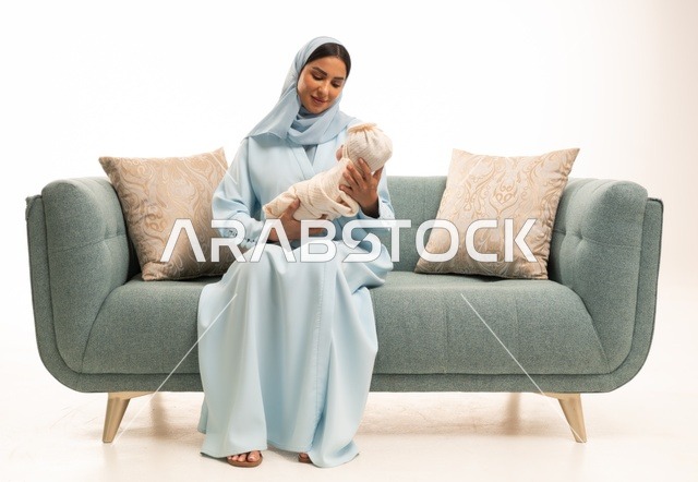 مساعدة الرضع على الاسترخاء والهدوء والنوم، تقوية رابط الأمومة بين الأم وطفلها، مفهوم البراءة والطفولة، تحمل المسؤولية والعناية بالأطفال،  بورتريه لإمرأة عربية خليجية سعودية محجبة تجلس على الاريكة تحمل صغيرها بإيماءات الحب والحنان, خلفية بيضاء