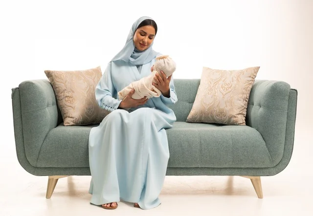 مساعدة الرضع على الاسترخاء والهدوء والنوم، تقوية رابط الأمومة بين الأم وطفلها، مفهوم البراءة والطفولة، تحمل المسؤولية والعناية بالأطفال،  بورتريه لإمرأة عربية خليجية سعودية محجبة تجلس على الاريكة تحمل صغيرها بإيماءات الحب والحنان, خلفية بيضاء