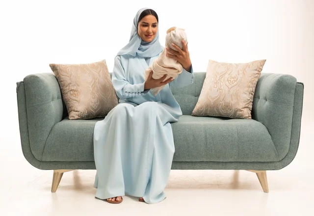 Helping infants relax, calm down, and sleep, strengthening the maternal bond between mother and child, the concept of innocence and childhood, taking responsibility and caring for children, a portrait of a Saudi Arabian Gulf woman in a hijab sitting on a sofa holding her baby with gestures of love and tenderness, white background.