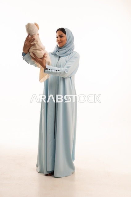 تقوية رابط الأمومة بين الأم وطفلها، مفهوم البراءة والطفولة، تحمل المسؤولية والعناية بالأطفال،  بورتريه لإمرأة عربية خليجية سعودية محجبة تحمل صغيرها بإيماءات الحب والحنان, الجسم بالطول الكامل، خلفية بيضاء