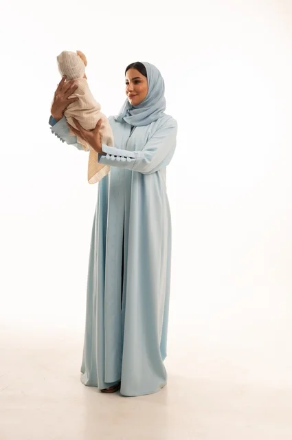 تقوية رابط الأمومة بين الأم وطفلها، مفهوم البراءة والطفولة، تحمل المسؤولية والعناية بالأطفال،  بورتريه لإمرأة عربية خليجية سعودية محجبة تحمل صغيرها بإيماءات الحب والحنان, الجسم بالطول الكامل، خلفية بيضاء
