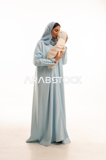 تقوية رابط الأمومة بين الأم وطفلها، مفهوم البراءة والطفولة، تحمل المسؤولية والعناية بالأطفال،  بورتريه لإمرأة عربية خليجية سعودية محجبة تحمل صغيرها بإيماءات الحب والحنان, الجسم بالطول الكامل، خلفية بيضاء