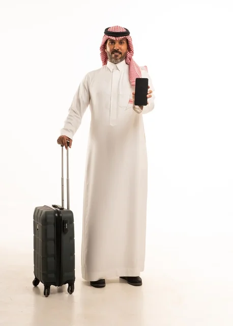 استخدام الأجهزة الحديثة و المتطورة , التجهيز للسفر والرحلات الممتعة, بورتريه لرجل عربي خليجي سعودي يرتدي الثوب التقليدي والشماغ يمسك بيده حقيبة السفر والهاتف المحمول, صورة للجسم بالطول الكامل , خلفية بيضاء