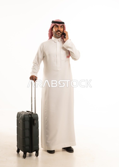 استخدام الأجهزة الحديثة و المتطورة , التجهيز للسفر والرحلات الممتعة, بورتريه لرجل عربي خليجي سعودي يرتدي الثوب التقليدي والشماغ يمسك بيده حقيبة السفر والهاتف المحمول, صورة للجسم بالطول الكامل , خلفية بيضاء