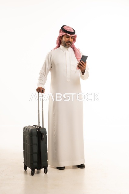 استخدام الأجهزة الحديثة و المتطورة , التجهيز للسفر والرحلات الممتعة, بورتريه لرجل عربي خليجي سعودي يرتدي الثوب التقليدي والشماغ يمسك بيده حقيبة السفر والهاتف المحمول, صورة للجسم بالطول الكامل , خلفية بيضاء