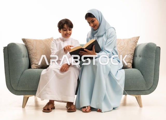 Reading beneficial stories for children, the concept of affection and tenderness among mothers, raising children and caring for them, strengthening the bonds of love and family cohesion, a portrait of a Saudi Gulf Arab mother in a hijab sitting on the sofa with her son reading an entertaining story, gestures of happiness and joy, white background.