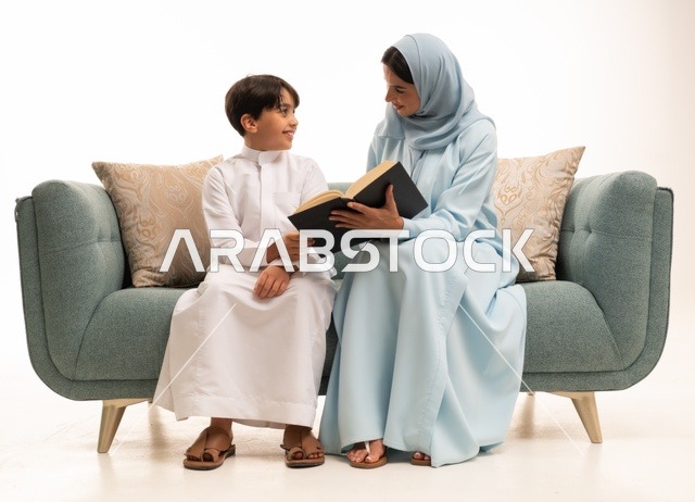 قراءه القصص المفيده للأبناء، مفهوم العطف والحنان عند الأمهات، تربية الأبناء والاهتمام بهم، تقوية روابط المحبة والتماسك الأسري، بورتريه لأم عربية خليجية سعودية محجبة تجلس على الاريكة برفقة ولدها تقوم بقراءة قصة مسلية، ايماءات السعادة والسرور، خلفية بيضاء