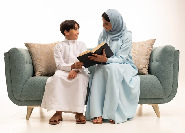 قراءه القصص المفيده للأبناء، مفهوم العطف والحنان عند الأمهات، تربية الأبناء والاهتمام بهم، تقوية روابط المحبة والتماسك الأسري، بورتريه لأم عربية خليجية سعودية محجبة تجلس على الاريكة برفقة ولدها تقوم بقراءة قصة مسلية، ايماءات السعادة والسرور، خلفية بيضاء