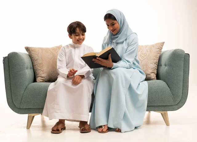قراءه القصص المفيده للأبناء، مفهوم العطف والحنان عند الأمهات، تربية الأبناء والاهتمام بهم، تقوية روابط المحبة والتماسك الأسري، بورتريه لأم عربية خليجية سعودية محجبة تجلس على الاريكة برفقة ولدها تقوم بقراءة قصة مسلية، ايماءات السعادة والسرور، خلفية بيضاء
