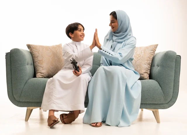 مفهوم الأسرة السعيدة، بورتريه لسيدة عربية خليجية سعودية تجلس في غرفة المعيشة برفقة ابنها، قضاء وقت ممتع بلعب ألعاب الفيديو بإيماءات تدل على السعادة ، ولد يمسك عصا التحكم وتلعب بألعاب الفيديو بجانب والدته ، المتعة و التسلية  بألعاب البلايستيشن الترفيهية ، خلفية بيضاء