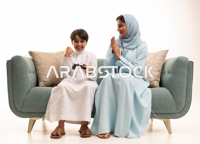 مفهوم الأسرة السعيدة، بورتريه لسيدة عربية خليجية سعودية تجلس في غرفة المعيشة برفقة ابنها، قضاء وقت ممتع بلعب ألعاب الفيديو بإيماءات تدل على السعادة ، ولد يمسك عصا التحكم وتلعب بألعاب الفيديو بجانب والدته ، المتعة و التسلية  بألعاب البلايستيشن الترفيهية ، خلفية بيضاء