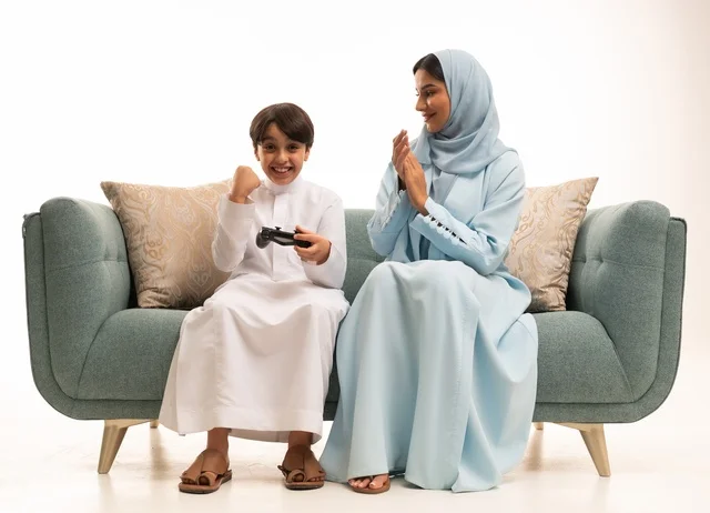 The concept of a happy family, a portrait of a Saudi Gulf Arab woman sitting in the living room with her son, enjoying time playing video games with gestures indicating happiness, a boy holding a controller and playing video games next to his mother, fun and entertainment with recreational PlayStation games, white background.
