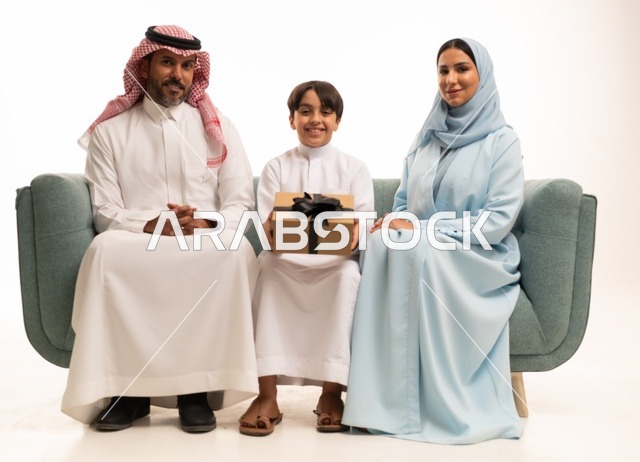 الألفة والمودة بين أفراد الأسرة السعودية, بورتريه  لعائلة عربية خليجية سعودية تجلس على الاريكة تقدم الهدية لولدها بايماءات الرضا والسرور, تقديم الهدايا بالمناسبات وأعياد الميلاد, خلفية بيضاء