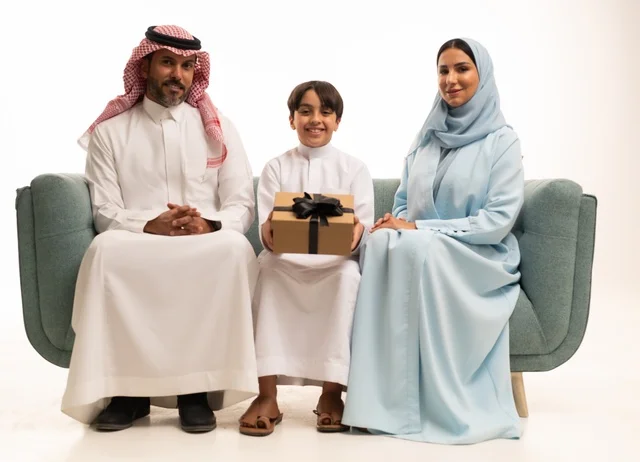 الألفة والمودة بين أفراد الأسرة السعودية, بورتريه  لعائلة عربية خليجية سعودية تجلس على الاريكة تقدم الهدية لولدها بايماءات الرضا والسرور, تقديم الهدايا بالمناسبات وأعياد الميلاد, خلفية بيضاء