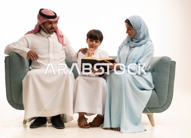 قراءة القصص المفيدية للابناء، الاهتمام بتعليم بالأبناء ودعمهم، الانشطة التعليمية وتنمية القدرات، بورتريه لزوجان عربيان خليجيان سعوديان يرتديان الزي التقليدي يجلسان على الاريكة يقومان بتعليم ابنهم، استغلال أوقات الفراغ بممارسة هواية المطالعة، خلفية بيضاء