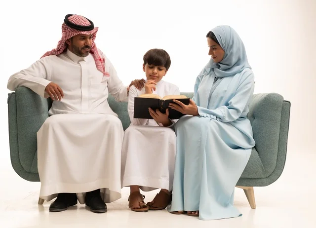 Reading beneficial stories for children, focusing on educating and supporting children, educational activities and developing skills, a portrait of an Arab Gulf couple from Saudi Arabia wearing traditional attire sitting on a sofa teaching their son, utilizing free time by engaging in the hobby of reading, white background.