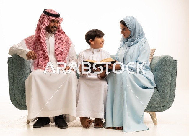 قراءة القصص المفيدية للابناء، الاهتمام بتعليم بالأبناء ودعمهم، الانشطة التعليمية وتنمية القدرات، بورتريه لزوجان عربيان خليجيان سعوديان يرتديان الزي التقليدي يجلسان على الاريكة يقومان بتعليم ابنهم، استغلال أوقات الفراغ بممارسة هواية المطالعة، خلفية بيضاء