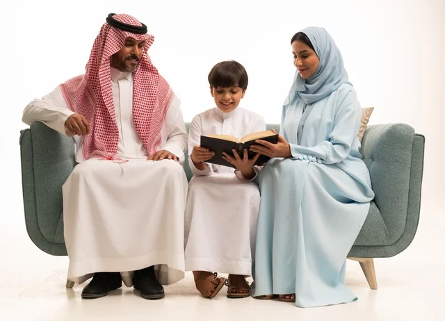 قراءة القصص المفيدية للابناء، الاهتمام بتعليم بالأبناء ودعمهم، الانشطة التعليمية وتنمية القدرات، بورتريه لزوجان عربيان خليجيان سعوديان يرتديان الزي التقليدي يجلسان على الاريكة يقومان بتعليم ابنهم، استغلال أوقات الفراغ بممارسة هواية المطالعة، خلفية بيضاء