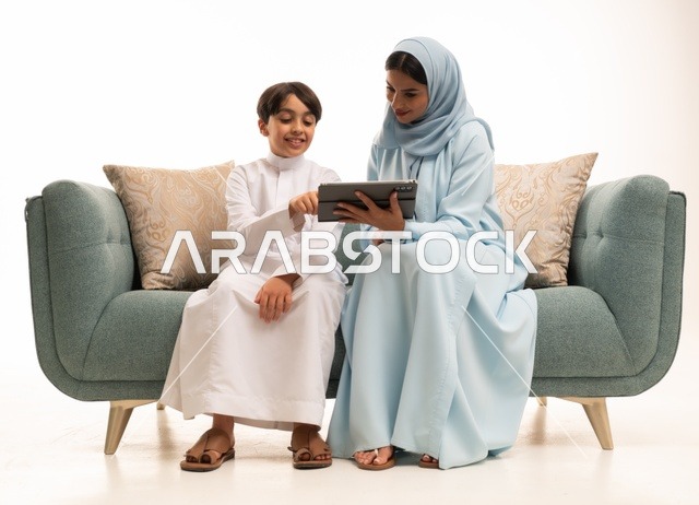 الأجواء العائلية المليئة بالحب والترابط والاستقرار، متابعة حصص الأونلاين وإجراء مكالمة فيديو من خلال التابليت المحمول، استخدام جهاز تقني حديث، بورتريه لولد عربي سعودي خليجي يرتدي الثوب التقليدي يستخدم الجهاز اللوحي ويجلس على الاريكة برفقة والدته، خلفية بيضاء