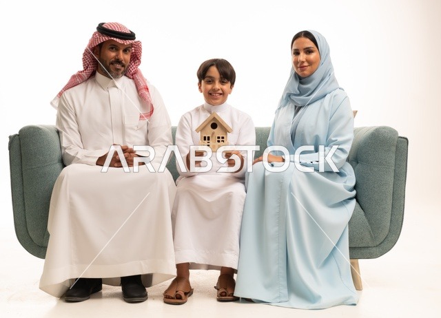 الحفاظ على الاستقرار الأسري والترابط العائلي، بورتريه  لولد عربي خليجي سعودي مبتسم ترتدي الزي التقليدي ويجلس على الاريكه امام والديه يحمل بيده مجسم صغير على شكل منزل، الاستمتاع بالسكن الجديد، خلفيه بيضاء