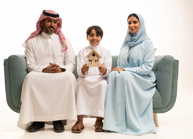 Maintaining family stability and family ties, a portrait of a smiling Arab Gulf Saudi boy wearing traditional attire sitting on a sofa in front of his parents holding a small model in the shape of a house, enjoying the new home, white background.
