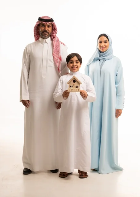 النظر إلى الكاميرا بإيماءات السرور، الحفاظ على الاستقرار الأسري والترابط العائلي، بورتريه  لولد عربي خليجي سعودي مبتسم ترتدي الزي التقليدي ويقف مع والديه يحمل بيده مجسم صغير على شكل منزل، الاستمتاع بالسكن الجديد، خلفيه بيضاء