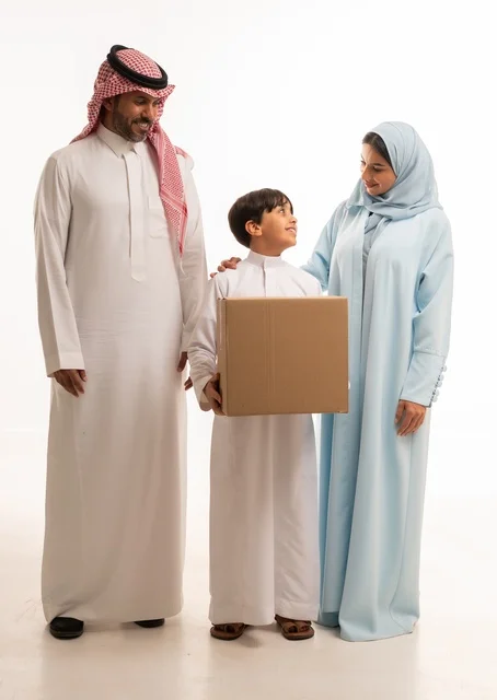 التعاون والتشارك في نقل صناديق الأغراض المنزلية، بورتريه لعائلة عربية خليجية سعودية يقفون في البيت الجديد ينظرون إلى الكاميرا بايماءات السعادة والسرور بامتلاك سكن جديد، ولد يحمل صندوق كرتوني، ترتيب وتنظيم المنزل، مفهوم الاسكان والتمليك والاستقرار العائلي، خلفية بيضاء