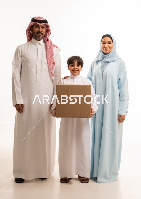 التعاون والتشارك في نقل صناديق الأغراض المنزلية، بورتريه لعائلة عربية خليجية سعودية يقفون في البيت الجديد ينظرون إلى الكاميرا بايماءات السعادة والسرور بامتلاك سكن جديد، ولد يحمل صندوق كرتوني، ترتيب وتنظيم المنزل، مفهوم الاسكان والتمليك والاستقرار العائلي، خلفية بيضاء