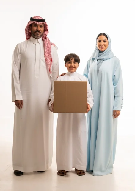 التعاون والتشارك في نقل صناديق الأغراض المنزلية، بورتريه لعائلة عربية خليجية سعودية يقفون في البيت الجديد ينظرون إلى الكاميرا بايماءات السعادة والسرور بامتلاك سكن جديد، ولد يحمل صندوق كرتوني، ترتيب وتنظيم المنزل، مفهوم الاسكان والتمليك والاستقرار العائلي، خلفية بيضاء