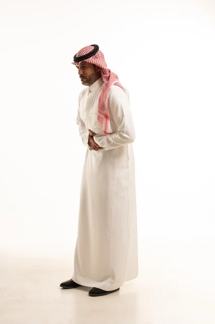 تعابير يد على البطن وجود خطبا ما، الاهتمام بالمظهر الخارجي والأناقة، تعابيرالألم والمعاناة، بورتريه لرجل عربي خليجي سعودي يرتدي الثوب التقليدي، شاب سعودي ،مفهوم التعبير بالإشارة، صورة الجسم بالطول الكامل، خلفية بيضاء