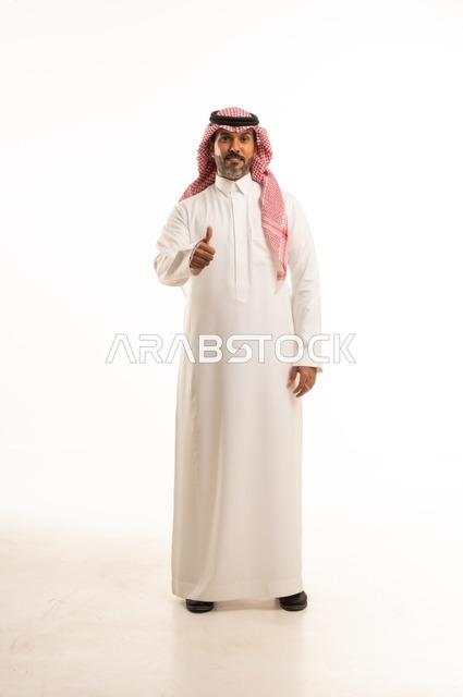 أشارة باستخدام اصبع الأبهام للأعلى تعابير وجه ابتسامة، الاهتمام بالمظهر الخارجي والأناقة، تعابير السرور والسعادة، بورتريه لرجل عربي خليجي سعودي يرتدي الثوب التقليدي، شاب سعودي ،مفهوم التعبير بالإشارة، صورة الجسم بالطول الكامل، خلفية بيضاء