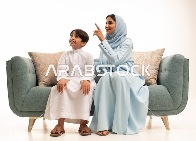 الإشارة بإصبع السبابة إلى شيء ما، قضاء أوقات عائليه سعيده في أيام العطله، نقاشات وحوارات الامهات مع الأبناء، بورتريه لأم عربيه خليجيه سعودية تجلس على الاريكة وتتبادل أطراف الأحاديث مع ابنها، مفهوم العائله السعوديه المترابطه، بناء علاقات أسريه قويه، خلفية بيضاء.