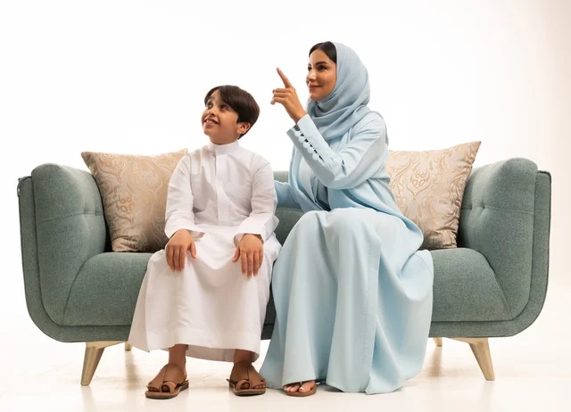 Pointing with the index finger at something, spending happy family times during holidays, discussions and dialogues between mothers and their children, a portrait of a Saudi Gulf Arab mother sitting on the sofa and exchanging conversations with her son, the concept of the interconnected Saudi family, building strong family relationships, white background.