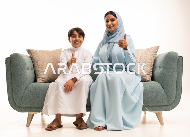 النظر إلى الكاميرا والإشارة بإصبع الابهام بإيماءات الاعجاب، قضاء أوقات عائليه سعيده في أيام العطله، نقاشات وحوارات الامهات مع الأبناء، بورتريه لأم عربيه خليجيه سعودية تجلس على الاريكة مع ابنها، مفهوم العائله السعوديه المترابطه، بناء علاقات أسريه قويه، خلفية بيضاء.