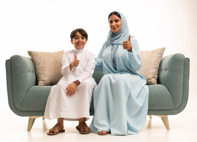 النظر إلى الكاميرا والإشارة بإصبع الابهام بإيماءات الاعجاب، قضاء أوقات عائليه سعيده في أيام العطله، نقاشات وحوارات الامهات مع الأبناء، بورتريه لأم عربيه خليجيه سعودية تجلس على الاريكة مع ابنها، مفهوم العائله السعوديه المترابطه، بناء علاقات أسريه قويه، خلفية بيضاء.