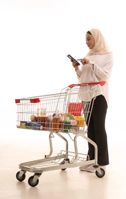 الاستفادة من الخصومات والتنزيلات على المنتجات، استخدام الهاتف المحمول أثناء التسوق في المولات والمراكز التجارية، بورتريه لشابة عربيه خليجية سعودية محجبة ترتدي الزي الكاجول تحمل الجوال بيدها وأمامها عربة المشتريات، الجسم بالطول الكامل، خلفية بيضاء