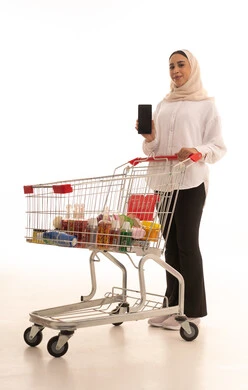 عرض شاشة هاتف محمول سوداء فارغة، الاستمتاع بالتسوق في المولات والمراكز التجارية، الاستفادة من الخصومات والتنزيلات، بورتريه لشابة عربية خليجية سعودية محجبة الزي الكاجوال تقف وتمسك الجوال بيدها وأمامها عربة المشتريات، الجسم بالطول الكامل، خلفية بيضاء