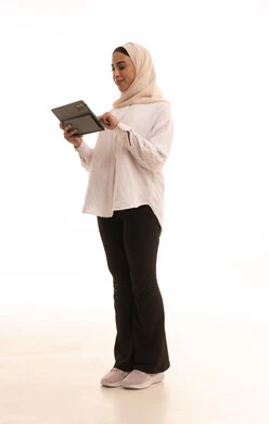 تصفح مواقع ووسائل التواصل الاجتماعي، مفهوم انجار المشاريع عن بعد، دمج التقنية في الحياة اليومية والعملية، بورتريه من الجانب لشابة عربيه خليجية سعودية محجبة ترتدي الزي الكاجول تستخدم جهاز التابلت، الجسم بالطول الكامل، خلفية بيضاء