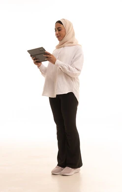 The concept of remote project completion, browsing websites and social media, integrating technology into daily and professional life, a side portrait of a young Arab Gulf Saudi woman wearing a hijab in casual attire using a tablet, full body, white background.