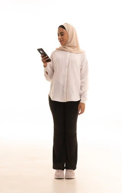 Browsing social media, having a good time on the phone with friends, a portrait of a young Arab Gulf Saudi woman wearing a hijab in casual attire sending text messages via mobile phone, full body, white background.