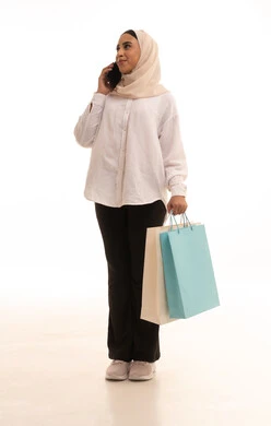 إجراء مكالمة هاتفية, الاستفادة من العروض والتخفيضات الموسمية, استخدام التكنولوجيا والتقنية المتطورة, بورتريه لشابة عربيه خليجية سعودية محجبة ترتدي الزي الكاجول تحمل بيدها الأكياس الورقية الملونة تمسك بيديها الهاتف المحمول, الجسم بالطول الكامل، خلفية بيضاء