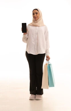 عرض شاشة سوداء فارغه عبر الجوال، الاستفادة من العروض والتخفيضات الموسمية, استخدام التكنولوجيا والتقنية المتطورة, بورتريه لشابة عربيه خليجية سعودية محجبة ترتدي الزي الكاجول تحمل بيدها الأكياس الورقية الملونة تمسك بيديها الهاتف المحمول, الجسم بالطول الكامل، خلفية بيضاء