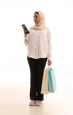 تصفح مواقع التواصل الاجتماعي عبر الجوال، الاستفادة من العروض والتخفيضات الموسمية, استخدام التكنولوجيا والتقنية المتطورة, بورتريه لشابة عربيه خليجية سعودية محجبة ترتدي الزي الكاجول تحمل بيدها الأكياس الورقية الملونة تمسك بيديها الهاتف المحمول, الجسم بالطول الكامل، خلفية بيضاء