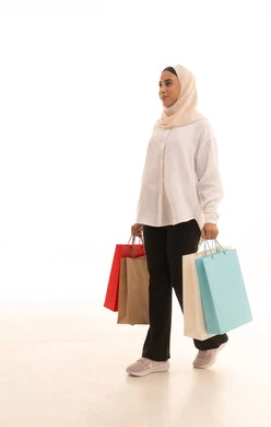 Taking advantage of seasonal offers and discounts, gestures of happiness and the feeling of enjoyment in shopping, buying gifts and necessities, the positive impact of shopping on women's psychology, a side portrait of a young Arab Gulf Saudi woman wearing a hijab in casual attire holding colorful shopping bags in her hand, full-body shot, white background.